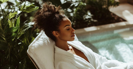 Black woman in a white robe sitting in a sunlit room with lush plants, practicing intentional rest and self-care.