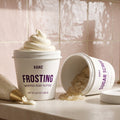 Kaike Frosting Whipped Body Butter in a white tub with a piped swirl of cream product on top, shown on a countertop with an additional piped bag, an open tub spilling sugar scrub, and a gold faucet in the background.